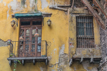 Old and decaying outer city wall, with a balcony with french wooden doors and iron railing, a double wooden window with iron bars, and yellow chipping paint, in Guanajuato, Mexico