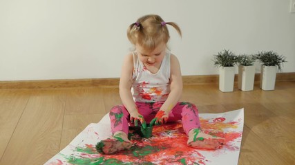 Little girl with smudgy paint fingers draws on a large sheet of white paper sitting on the floor