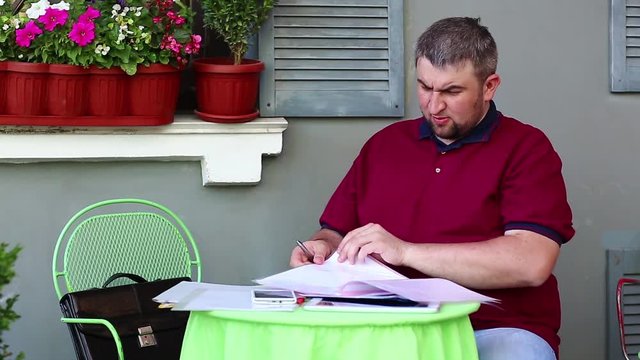 Funny man works with business papers. Businessman sits at the table in restaurant and holds his documents. Man sits in summer cafe, works with business papers and speaks to himself