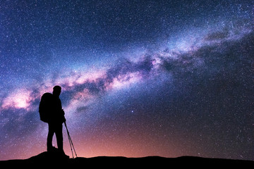 Silhouette of man with backpack and trekking poles against amazing purple Milky Way at night....