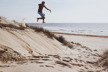 Young male jumping off a sandy dune on to the beach towards the water.