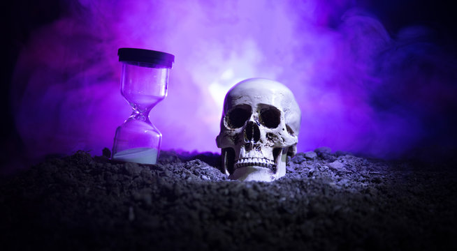 Skull and vintage hourglass on dark toned foggy background under beam of light. Horror concept.