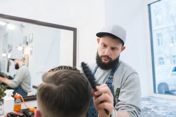 Barber with a beard creates a stylish hairstyle for a young man in a barbershop. A serious hairdresser cuts a man in a men's beauty salon