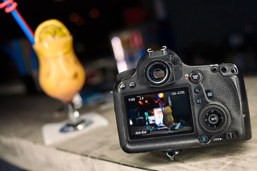 Professional modern DSLR camera low key image. Blurred yellow cocktail on background. Bar counter