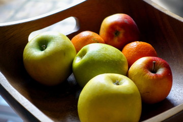 Square wooden fruit bowl containing imperfect red and green apples, and oranges