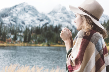 Adventure woman among mountains, enjoy the nature and feeling cold. Forest and lake wearing hat and poncho, boho and wanderlust style