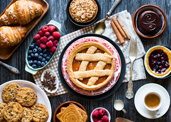 Top view of a wood table full of cakes, fruits, coffee, biscuits, spices and more