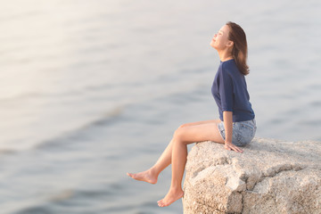 Side view portrait of Attractive fashion young Asian woman wearing casual sitting on the rock cliff relaxing, smiling and breathing deep fresh air with blurred sea wave ocean background.
