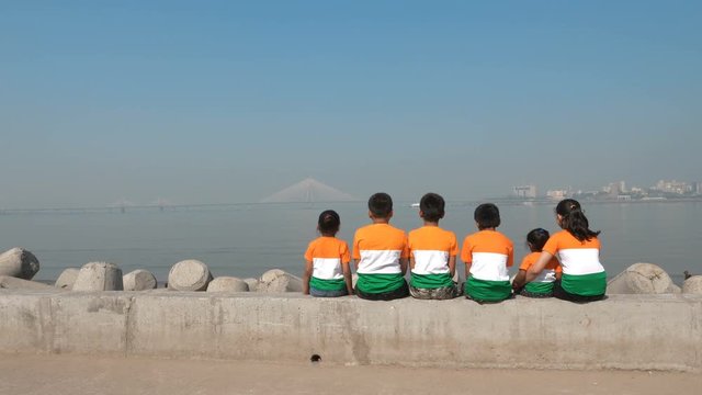 A group of children wearing Indian flag tricolor t-shirts sitting on a platform against sea and the modern bridge