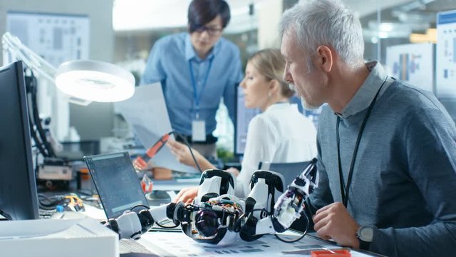 Senior Electronics Engineer Works With Robot, Programming it with Laptop. In the Background Laboratory with Technical Personnel Working.