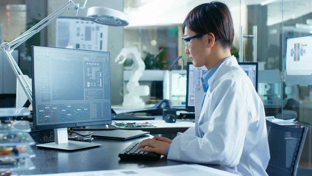 Asian Scientist Sitting at His Desk Doing Sophisticated Coding and Programming on His Desktop Computer. In the Background Computer Science Research Laboratory with Robotic Arm Model.