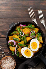 Warm potato salad with green beans, pepper, parsley, eggs and red onion in frying pan on old wooden background. Selective focus. Top view. Copy space.