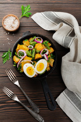 Warm potato salad with green beans, pepper, parsley, eggs and red onion in frying pan on old wooden background. Selective focus. Top view. Copy space.