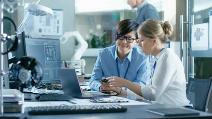 Asian Scientist Sitting at His Desk Consults Senior Engineer They Connect Printed Circuit Board to a Laptop and Start Coding. In the Background Computer Science Research Laboratory with Robotic Arm. - Powered by Adobe