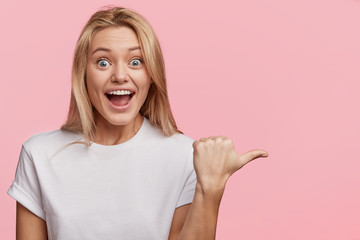 Attractive overjoyed female dressed casually, expresses surprisment and excitedness, indicates at...
