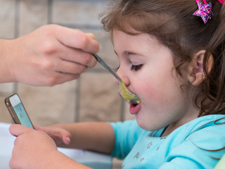 Mom feeding cute little girl while watching cartoons on mobile phone
