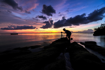 Silhouette of photographer taking photos of a dramatic sunset with tripod on the rock