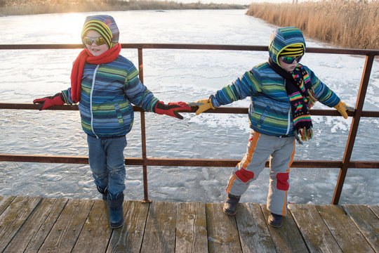 Happy boys in colorful winter clothes posing on a wooden bridge across a frozen river. Points for skiing, snowboarding and sledding. Kid playing outdoors in the snow. Outdoor fun for winter holiday