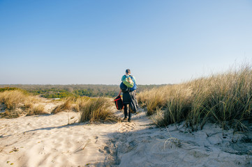 Homeless hermit carrying many bags with belongings in sandy dunes beyond sea in sunny day. Man from behind travelling alone. Unknown male with backpacks and heavy buggage in hands hiking outdoor.