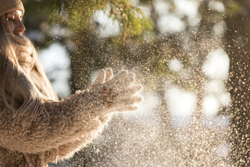 Close up of female hands in white knitted gloves, clap and shake hands out of snow at sunny winter day, snowflakes lightened by sun . Winter time concept.