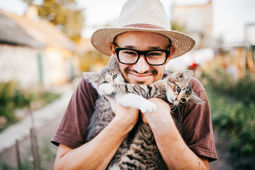 Happpy young bearded farmer holding two little kitten in hands outdoor in village with abstract background. Smiling man in glasses and straw hat playing with funny cute pets. Have fun in countryside. - Powered by Adobe