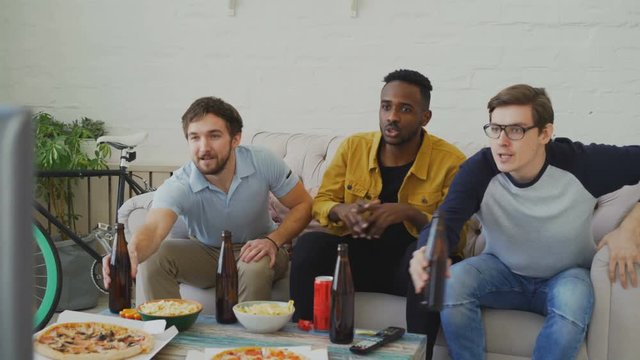 Group of young male friends watching sports match on TV together while drinking beer and eating snacks at home