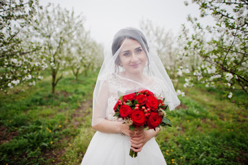 Portrait of a gentle young bride holding red bouquet in the blossoming garden on her wedding day.