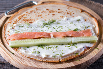 Pancake stuffed with cream cheese, dill, salmon and cucumber on a chopping wooden board