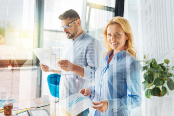 Cheerful woman. Young clever positive woman looking happy while standing not far from her concentrated colleague and smiling cheerfully