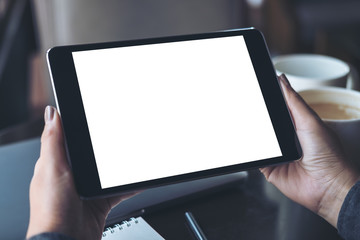 Mockup image of hands holding black tablet pc with blank white desktop screen with coffee cups on table in modern cafe