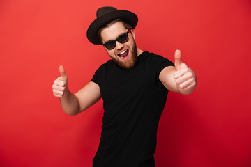 Image of excited young guy wearing black sunglasses and hat smiling and pointing fingers on camera...