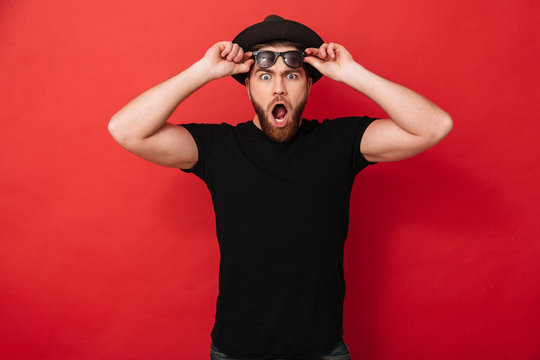 Portrait Of Surprised Stylish Man 30s In Hat Posing On Camera And Taking Off Black Sunglasses, Isolated Over Red Background