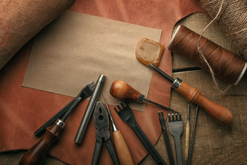 Naklejka premium Set of leather craft tools on wooden background. Workplace for shoemaker. Piece of hide and working handmade tools on a work table.