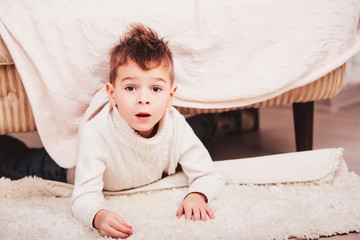 Fashionable guy in a white knitted sweater and a stylish haircut the Iroquois climbed out from under the couch and looks in the camera with his mouth open. Boy is lying on the floor under the sofa.