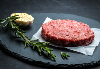 Raw burger beef patty with rosemary and garlic on dark cooking background