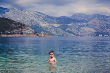Little smiling boy in the sea with blue and turquoise water. In the background blue mountains.