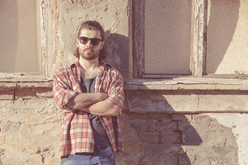 Man with beard and long hair posing in front of a ruined house.