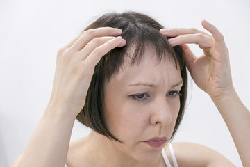 Naklejka premium Horrified young woman looking in the bathroom mirror staring open mouthed at the first grey hair on her scalp, a first sign of ageing, or noticing that she is suffering from dandruff