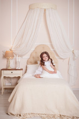 Fototapeta premium A little girl in her room sits on the bed and hugs a pillow. Child in white dress. Beige interior.