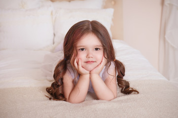 A little girl in her room, lying on the bed, leaning on her elbows. A large portrait. Child in white dress. Beige interior. © Надежда Манахова