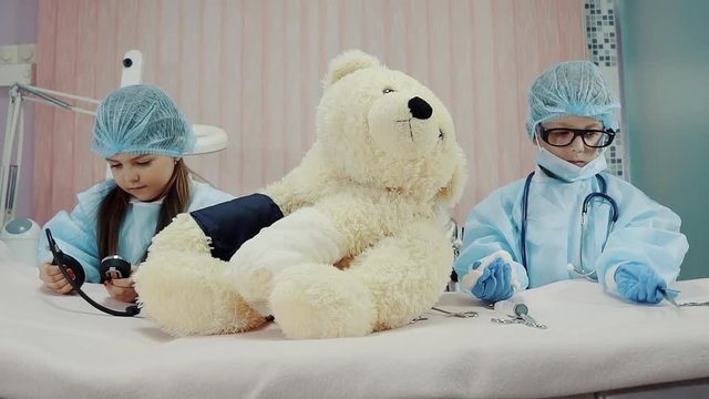 funny children in big gowns of doctors with a serious look measure the pulse on the paw of a teddy bear, the boy and the girl pretended to be doctors who work in a polyclinic