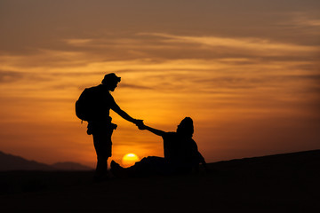 Silhouette of two men helping to stand up in desert of Sahara with beautiful sunset in background,...