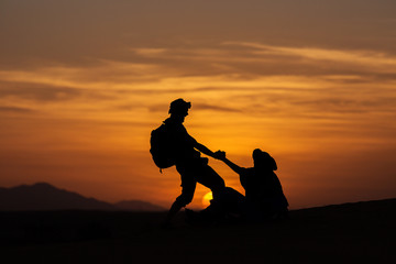 Silhouette of two men helping to stand up in desert of Sahara with beautiful sunset in background, Morocco