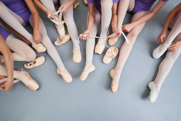 Obraz premium Little ballerinas putting on shoes, cropped image. Group of teen ballerinas sitting on the floor and adjusting their pointe shoes, top view.