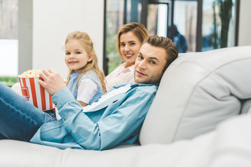 Fototapeta premium side view of family with popcorn looking at camera while watching film together at home
