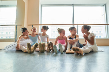 Fototapeta premium Young ballerinas sitting on floor and embracing. Cute little girls sitting on floor and laughing in ballet class together. They are best friends.
