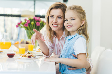 Obraz na płótnie Canvas smiling woman showing something mo daughter during breakfast at home