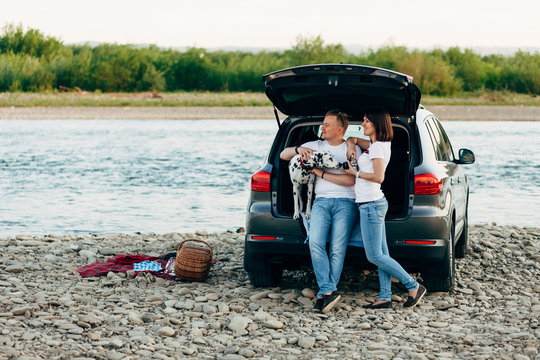 Portrait of happy young adult couple with dog on roadtrip. Man sitting on plaid with woman. Outdoor picnic concept.