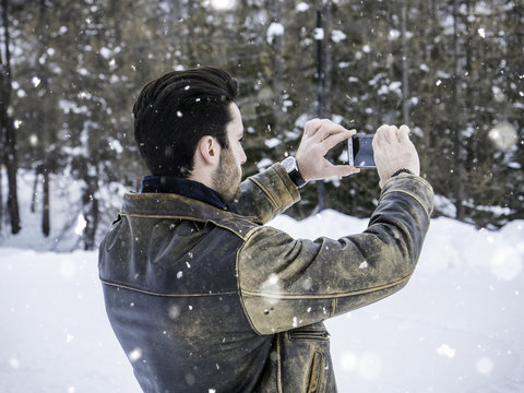 Handsome young man using smarpthone to take photographs of the landscape, while posing among snowy woods in the mountain