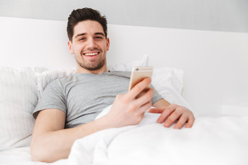 Photo of happy awake man in t-shirt smiling while lying alone in bed with white linen, and holding golden cell phone